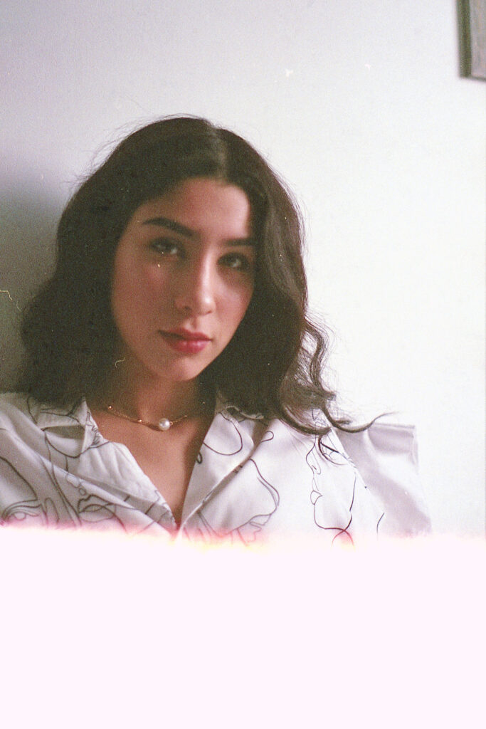 Nadya Inzunza Film Photo. A portrait of a brunette with wavy hair wearing a white blouse with abstract black lines set against a light-colored wall.